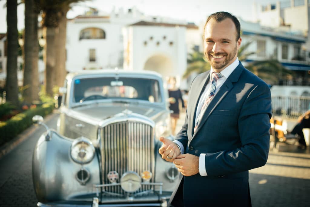 Man in a tailored navy suit smiling confidently while standing in front of a vintage silver luxury car in a sunny upscale location.